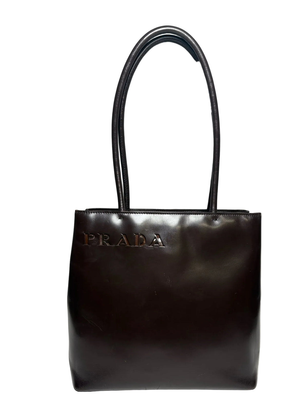 Brown leather Prada bag with two handles and an embossed "Prada" in the top left corner.  Bag is in front of a white background.