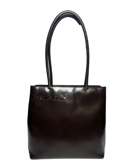 Brown leather Prada bag with two handles and an embossed "Prada" in the top left corner.  Bag is in front of a white background.